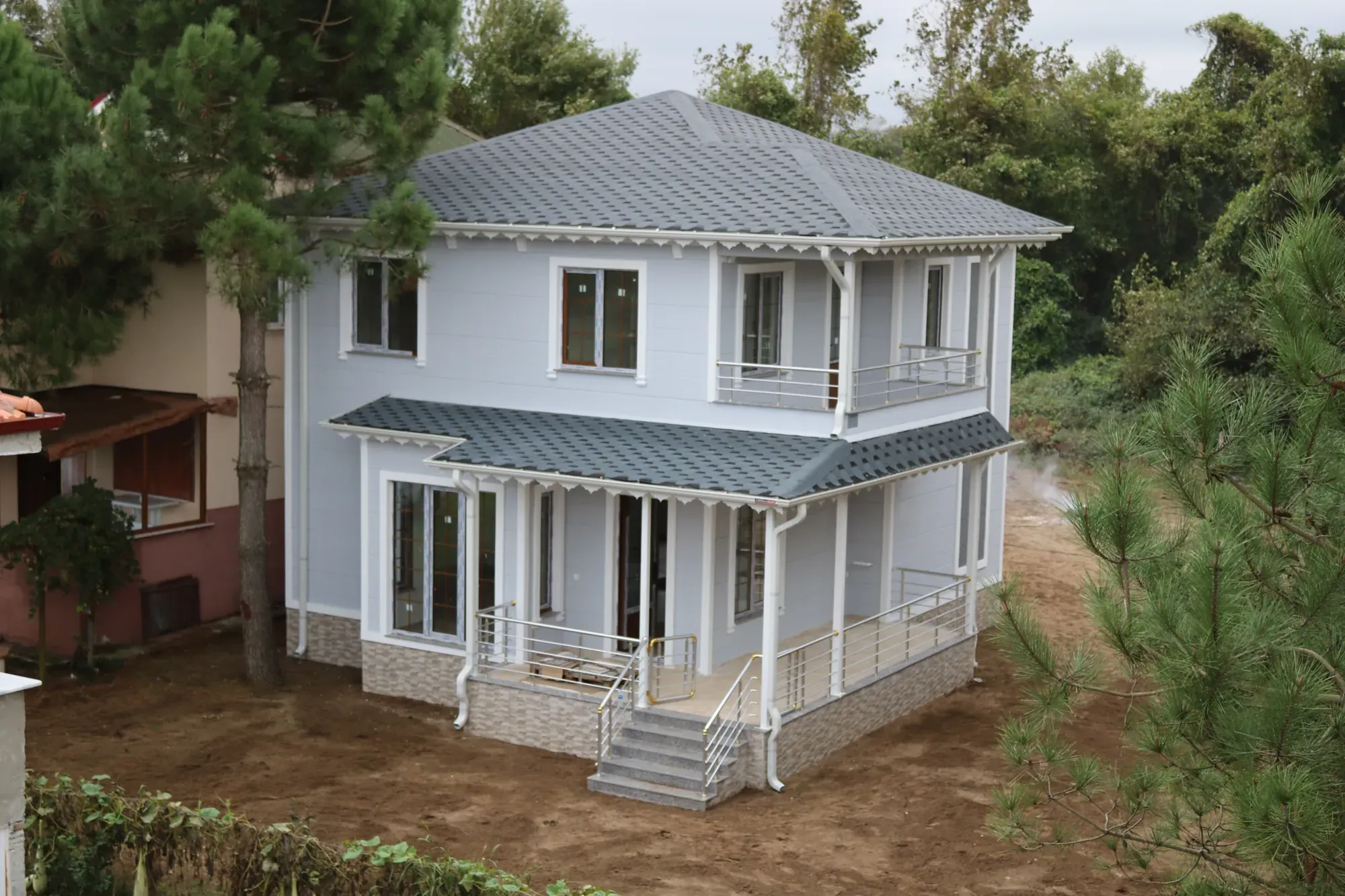 Çam ağaçları arasında beyaz cepheli, gri çatılı iki katlı 180 m² çelik konstrüksiyon ev