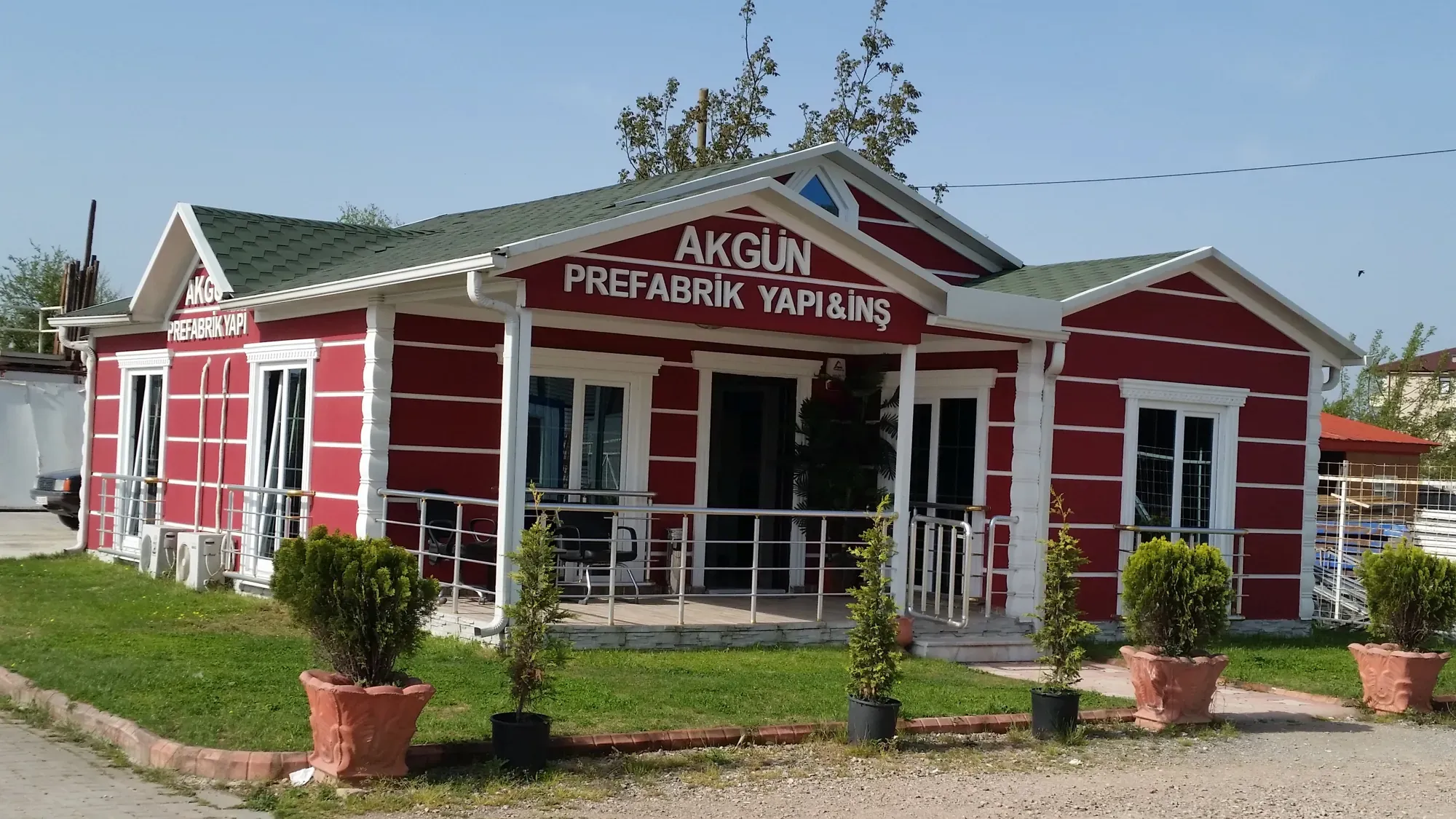 Alınlığında AKGÜN Prefabrik Yapı yazan kırmızı 95 m² prefabrik ev, önünde saksılı bitkiler
